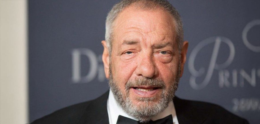 A man with short gray hair and a beard is wearing a black tuxedo and bow tie, standing in front of a blurred dark background with indistinct white text.