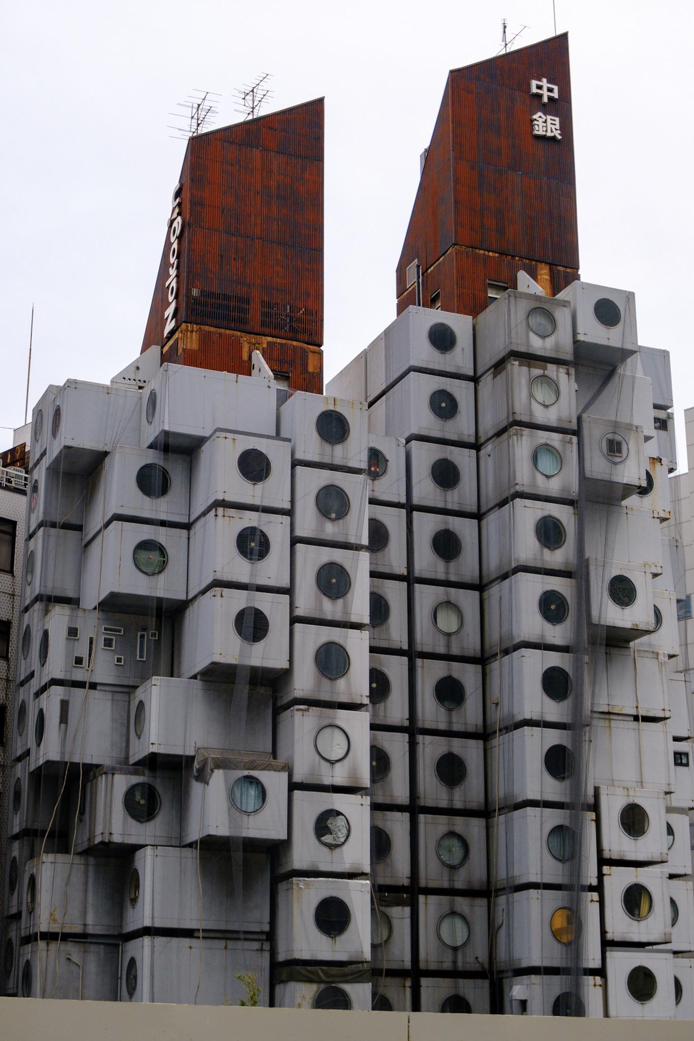 Futuristic building with stacked, cube-like modules featuring round windows; two tall, rust-colored structures with Japanese writing rise above the cubes. The sky is overcast.