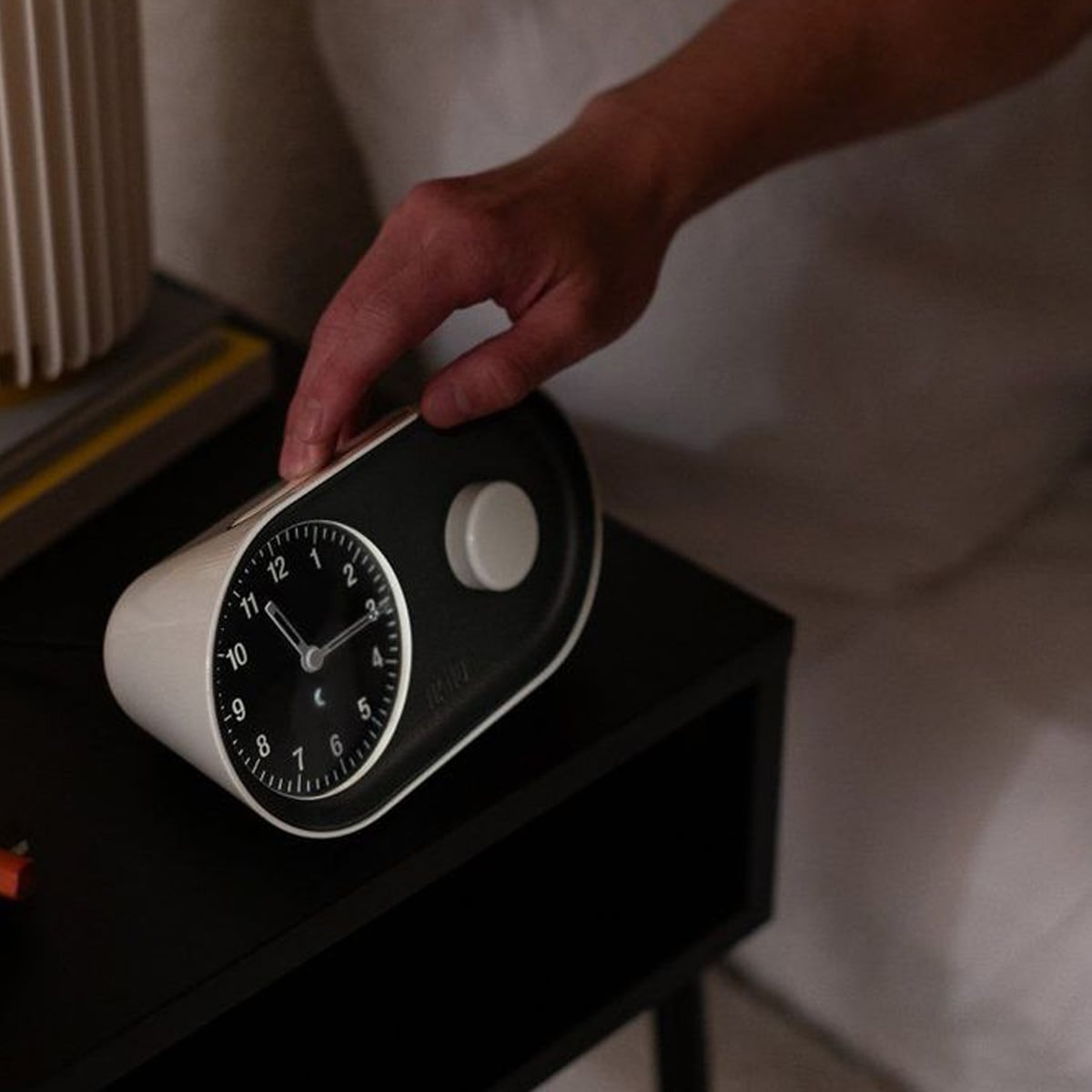 A hand reaches to turn off a black-and-white analog alarm clock on a bedside table, with part of a lamp and a bed visible in the background.