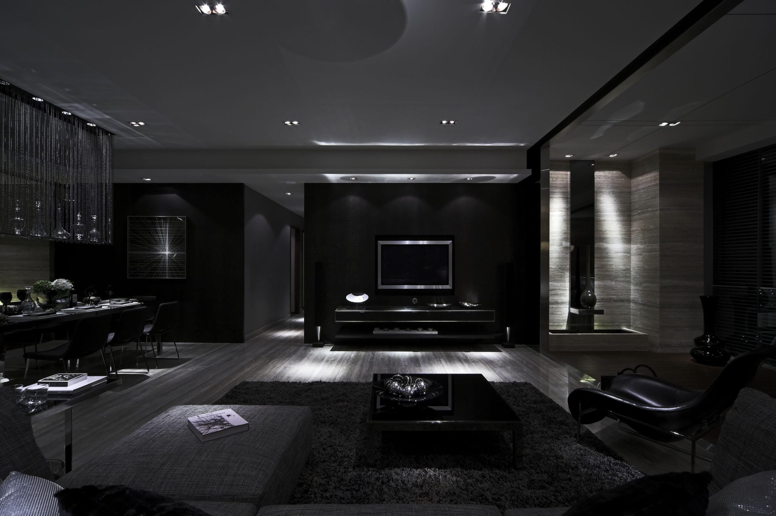 A modern living room with dark walls and decor, featuring a TV on a central wall, low furniture, a gray rug, a dining area on the left, and subtle ambient lighting throughout the space.