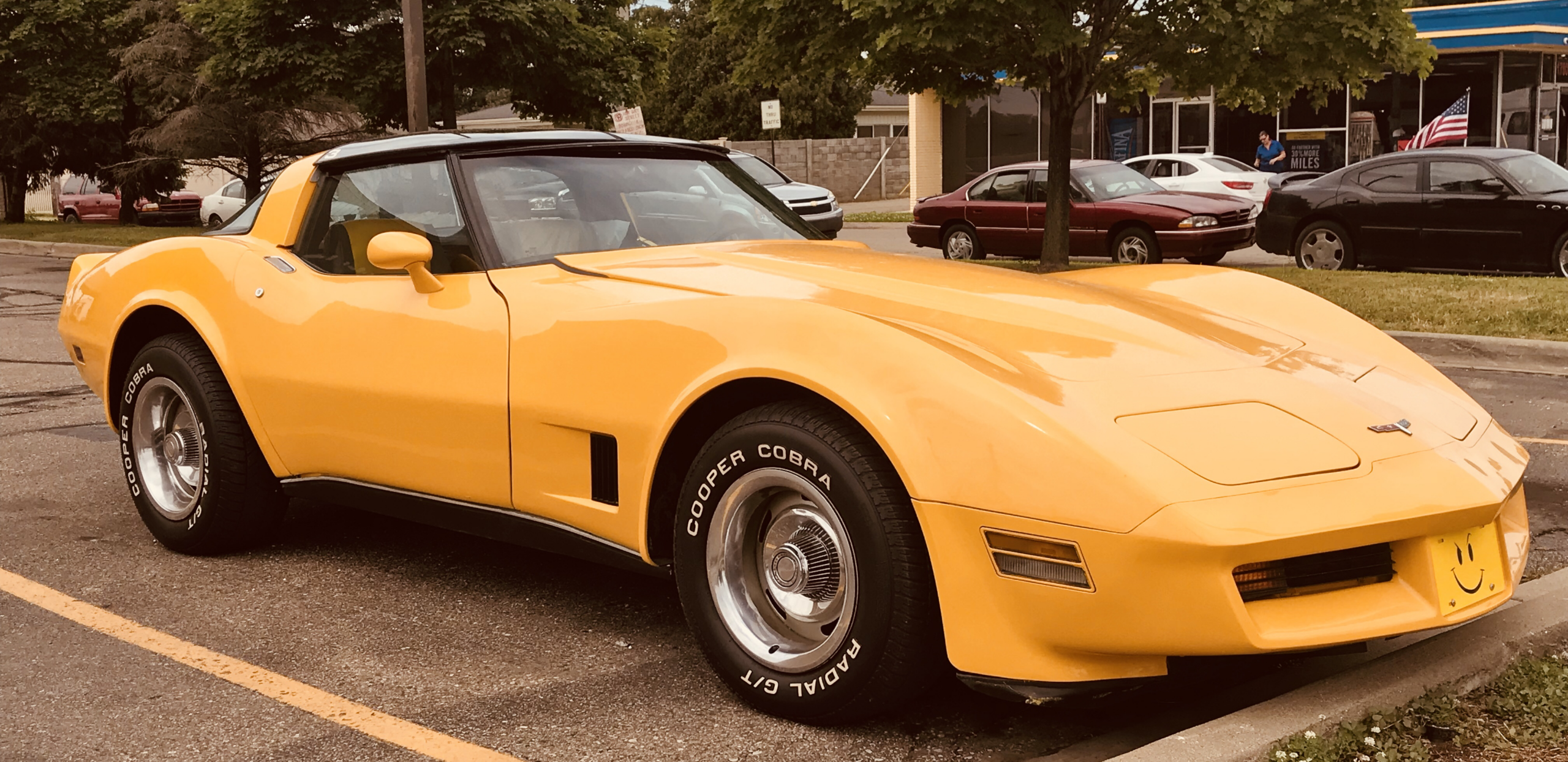 A yellow classic Chevrolet Corvette sports car with T-top roof is parked in a lot near a curb, surrounded by other parked cars and trees in the background.