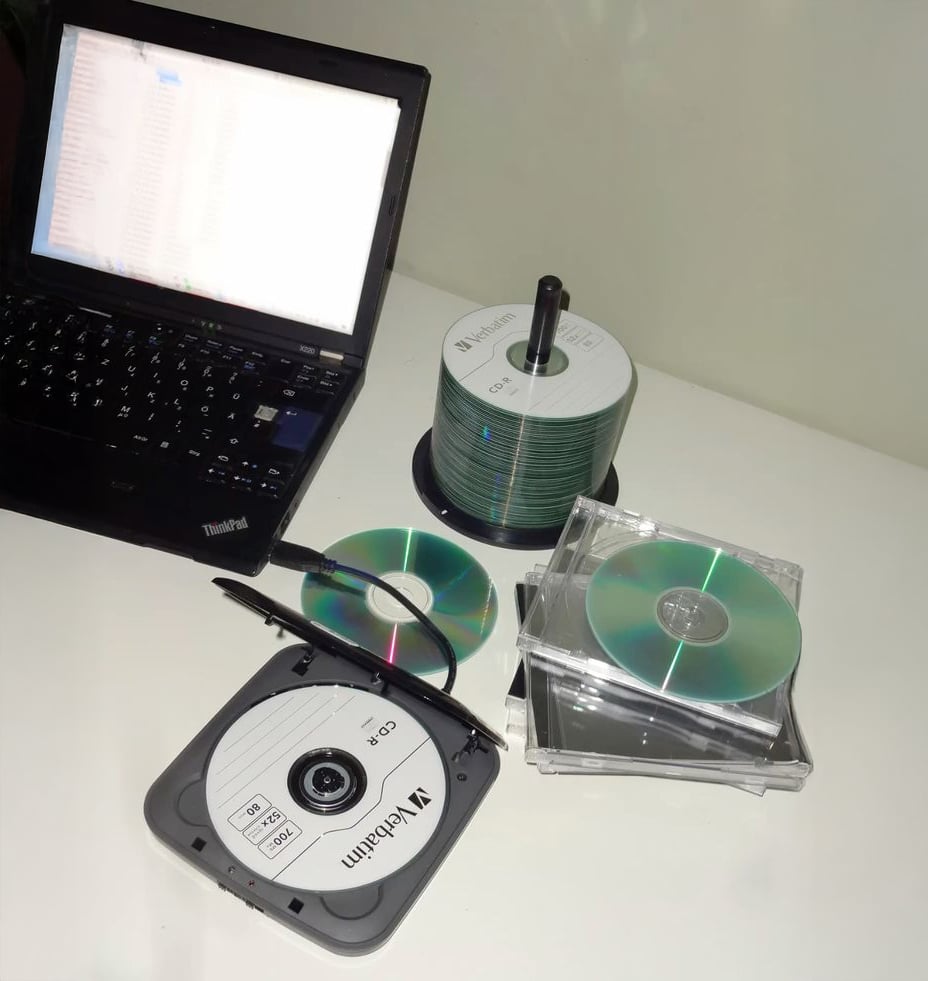 A laptop sits on a white desk next to a spindle of blank CDs, two CD cases, a marker, and a portable CD drive containing a labeled Verbatim CD-R. Two loose CDs are also on the desk.