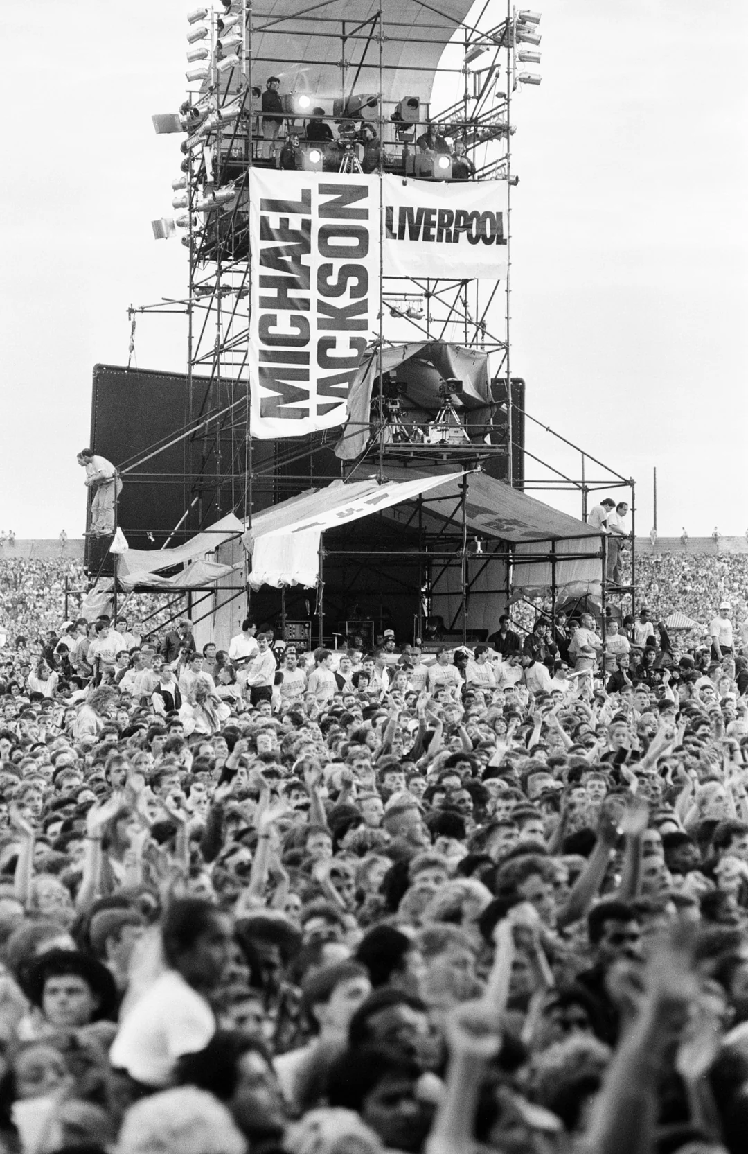 A large crowd gathers in front of a concert stage with banners reading "MICHAEL JACKSON" and "LIVERPOOL." People are cheering and raising their hands, and the stage is equipped with lights and speakers.