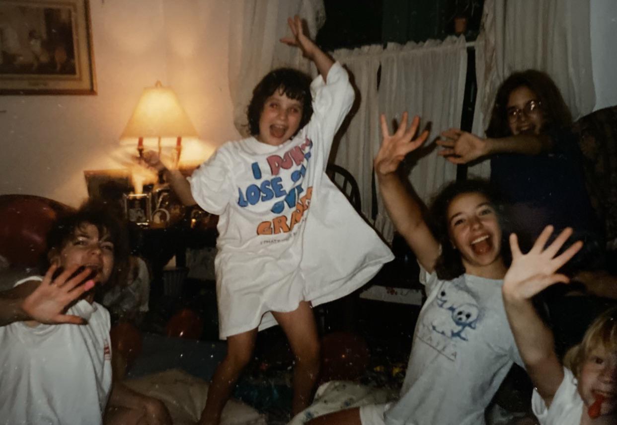 Four children in a living room joyfully pose with big smiles and raised hands. One child stands on a couch wearing an oversized T-shirt. The atmosphere is playful, and a lamp and curtains are visible in the background.