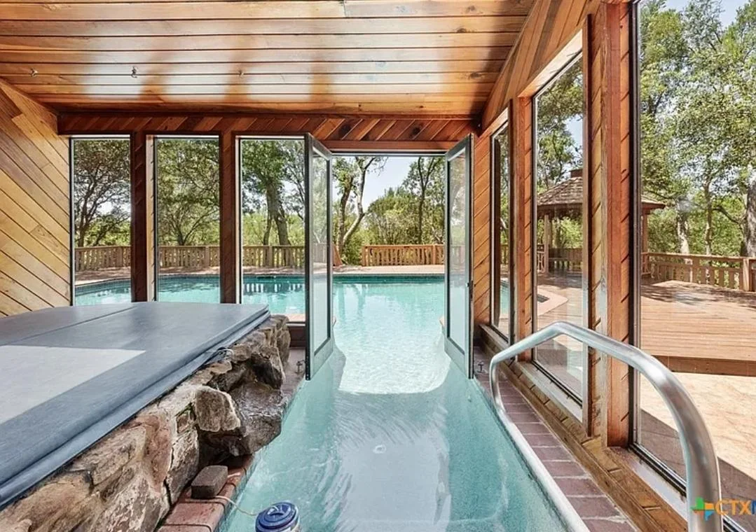 Indoor hot tub with wood-paneled walls opens to an outdoor pool through glass doors. Large windows provide a view of trees and a sunny wooden deck with a gazebo in the background.