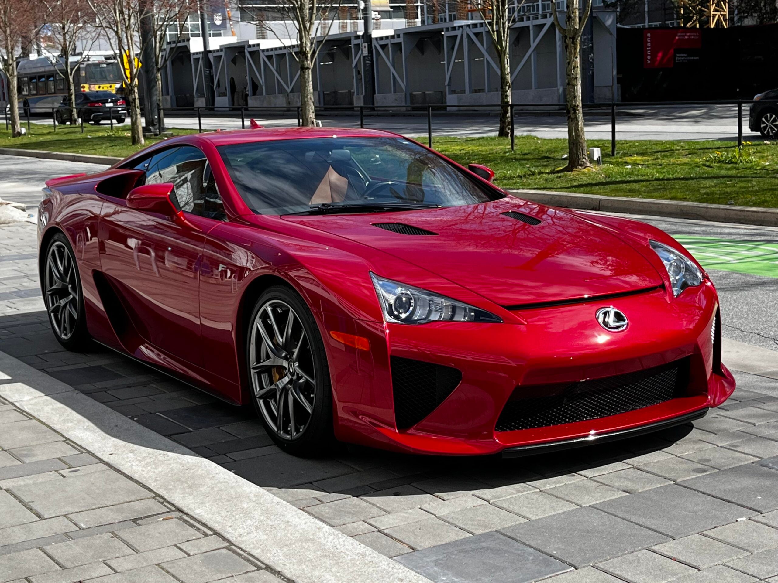 A red Lexus LFA sports car is parked on a paved street next to a sidewalk with trees and buildings in the background.