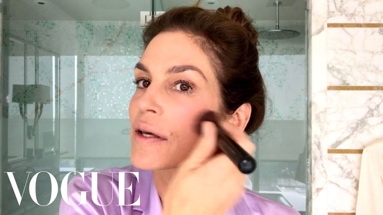 A woman wearing a lavender top applies makeup with a brush in a modern bathroom. The word "VOGUE" is overlaid in white text in the bottom left corner.