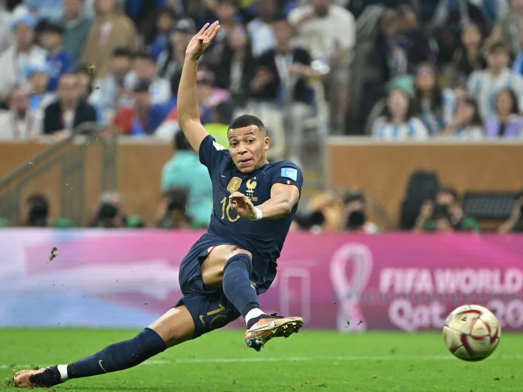 A soccer player in a blue France jersey is sliding on the field with one arm raised and the ball in front. The background shows spectators and a FIFA World Cup Qatar 2022 sign.