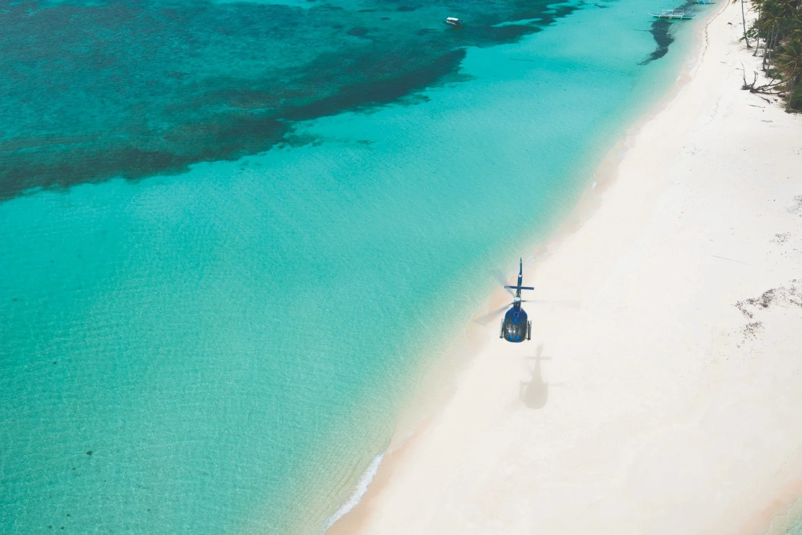A blue helicopter is parked on a pristine white sand beach next to clear turquoise water, with the ocean stretching into the distance and palm trees visible at the edge of the image.