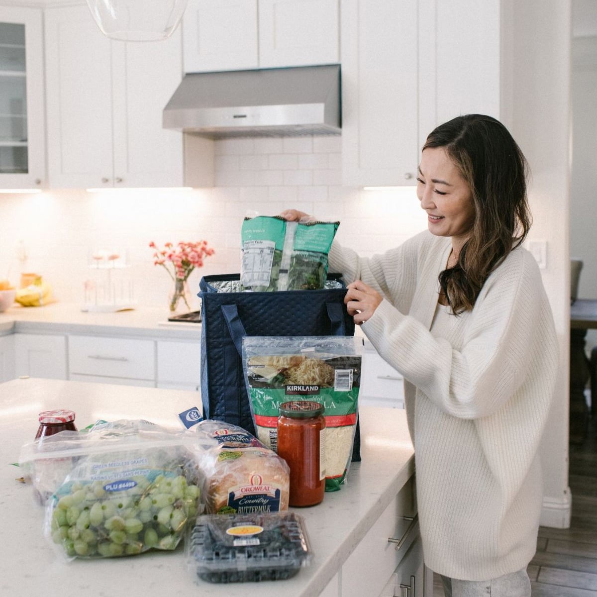 A woman in a white sweater smiles while unpacking groceries from a blue insulated bag on a white kitchen counter. Various food items, including bread, fruit, and greens, are spread out on the counter.