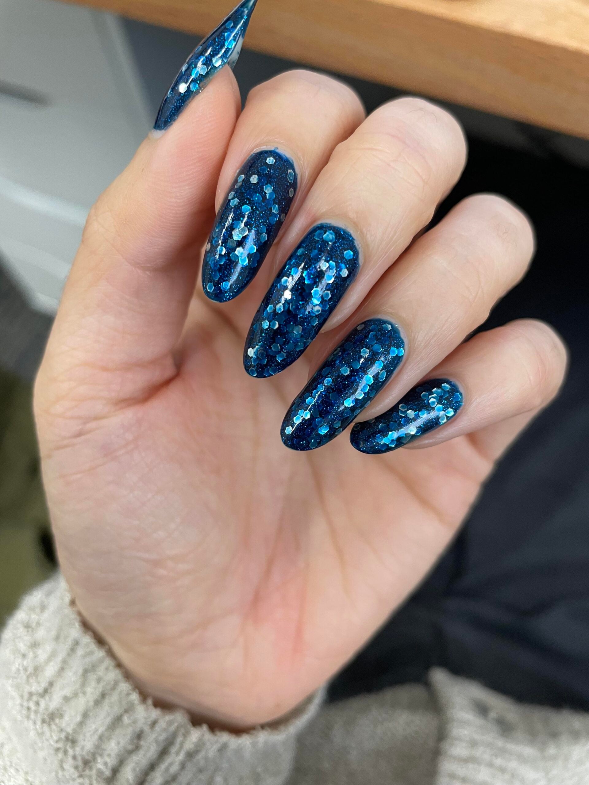 A hand with long, almond-shaped nails painted in dark blue polish with shiny blue and silver glitter. The person is wearing a light-colored sweater.