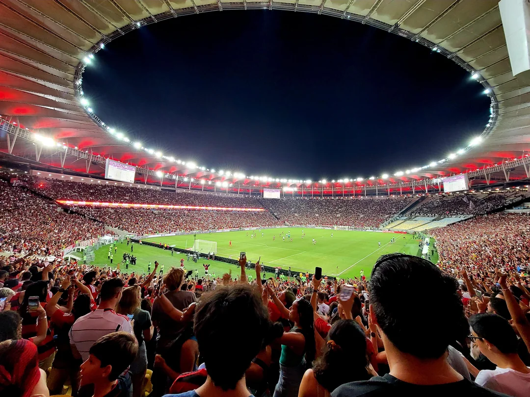 A large, packed stadium at night, filled with cheering fans watching a soccer match. Bright stadium lights and red accents illuminate the scene, while the crowd captures the moment with phones raised.