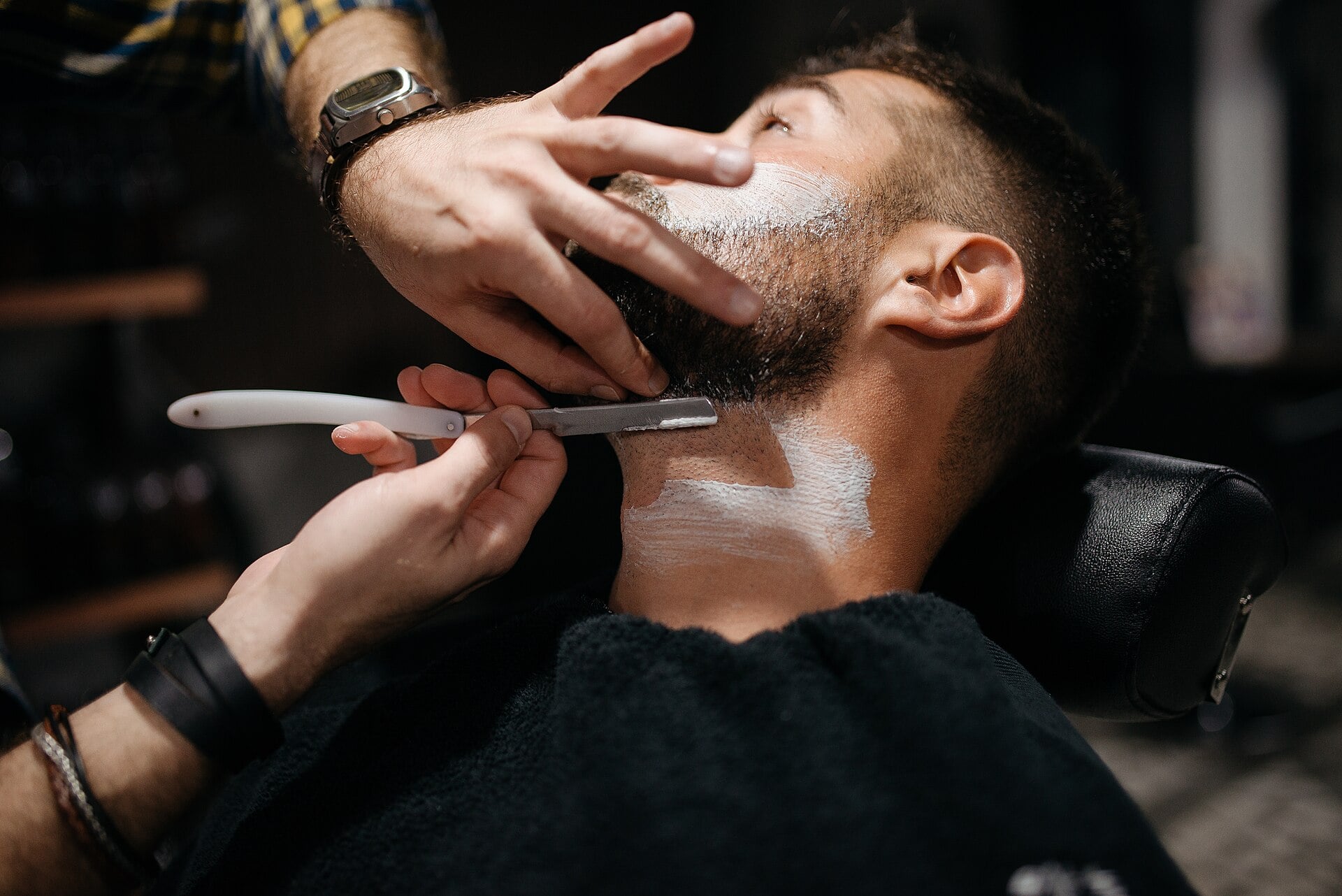 A man reclines in a barber chair while a barber uses a straight razor to shave his neck. The man has shaving cream applied and the barber’s hands gently hold his face and neck steady.