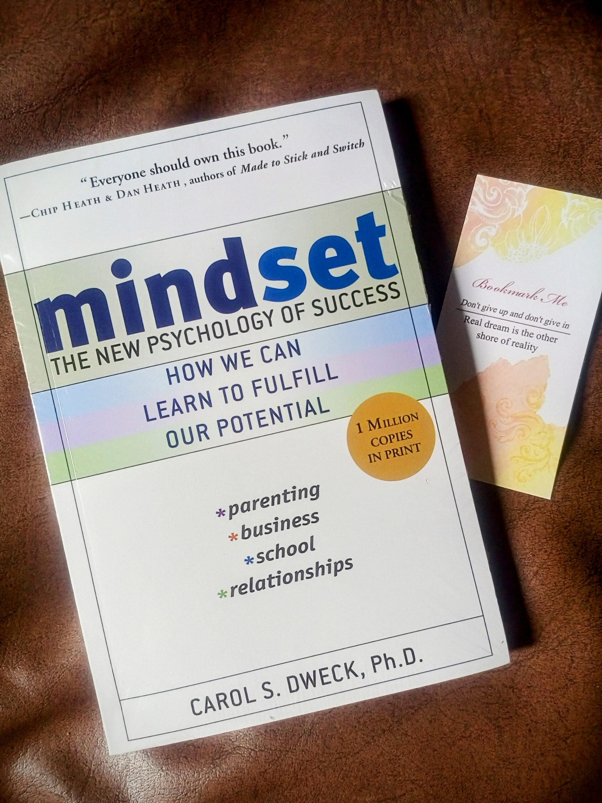A copy of the book "Mindset: The New Psychology of Success" by Carol S. Dweck lies on a brown surface next to a decorative card with an inspirational quote and soft yellow floral design.