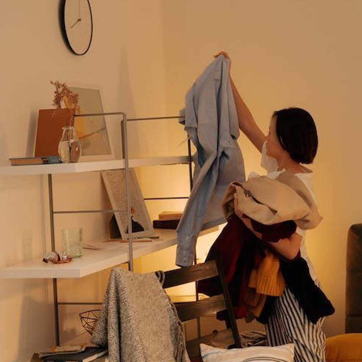 A person standing in a room holds several pieces of clothing and lifts a blue shirt while facing a shelf with books, decor, and a clock on the wall. The space appears warmly lit and slightly cluttered. Home.