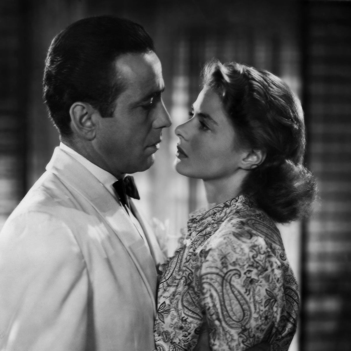 A man in a white suit and bow tie gazes intently at a woman with styled hair and a patterned dress as they stand close together, suggesting a dramatic or romantic moment in a classic black-and-white film scene.