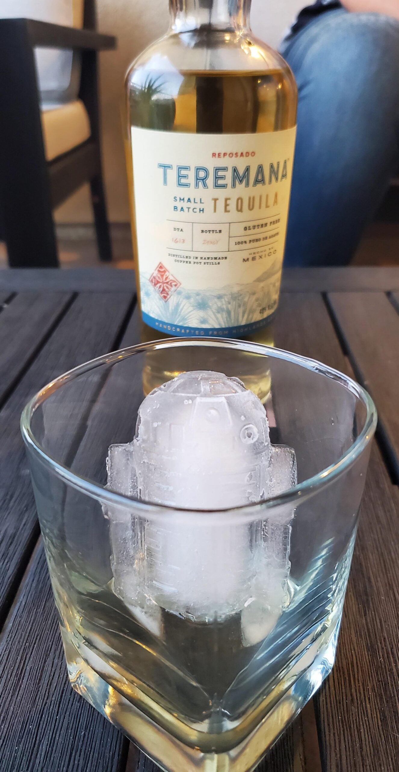 A glass with a large ice cube shaped like the Star Wars droid R2-D2 sits on a table, with a bottle of Teremana Reposado Tequila and a seated person in the background.