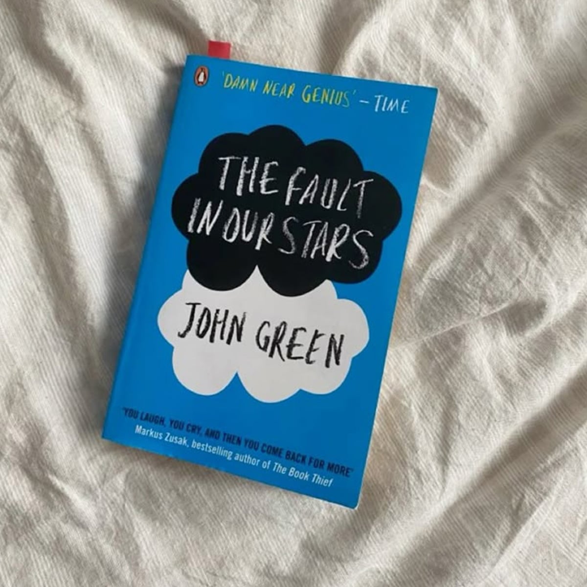 A paperback copy of "The Fault in Our Stars" by John Green with a bright blue cover rests on a textured, light-colored fabric surface.