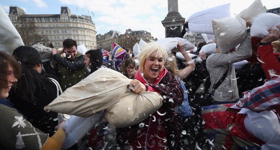 A group of people joyfully participate in a pillow fight outdoors, with feathers flying in the air. Smiling faces and energetic movement fill the scene in what appears to be a city square.