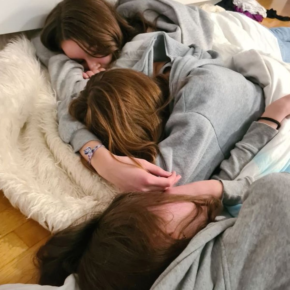 Three people with long hair wearing casual clothes are lying close together on the floor, appearing to be asleep or resting on a soft white rug and blanket. Their heads are nestled together, facing different directions.