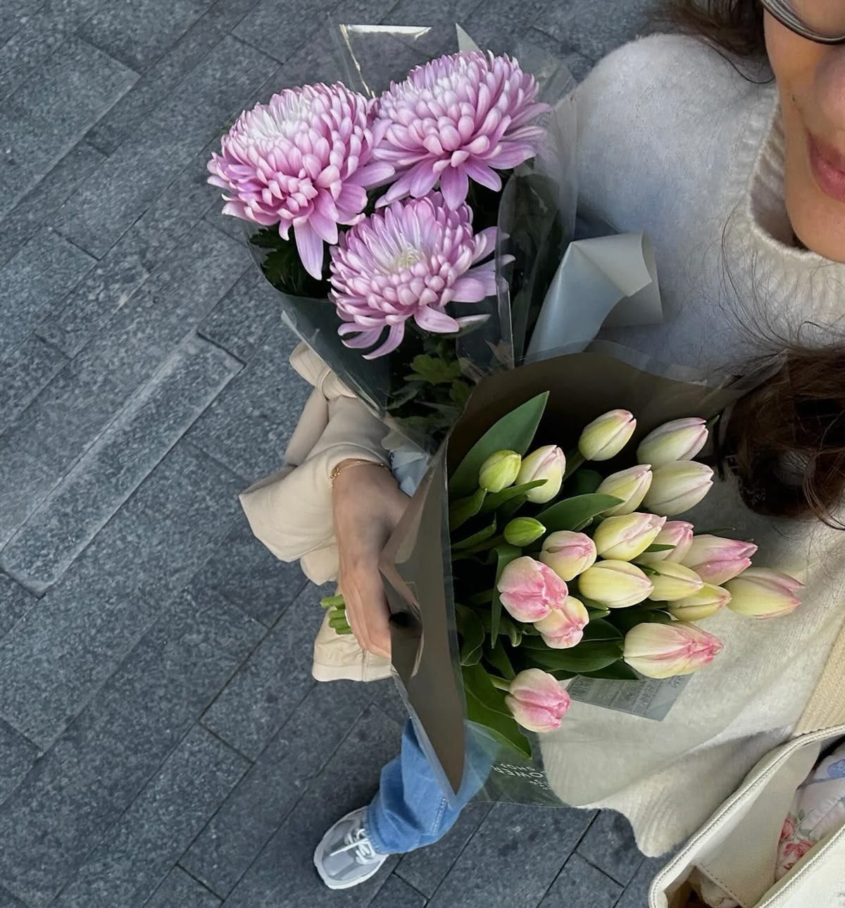 A person in a cream sweater and blue jeans holds two bouquets—one of pink chrysanthemums and one of pink and white tulips—while standing on a gray stone pavement. Only part of their face is visible.