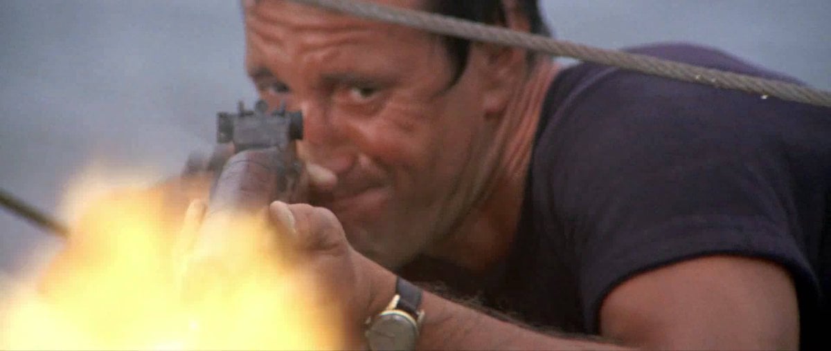 A man in a dark shirt aims and fires a rifle, with a bright muzzle flash visible. He has a determined expression and is outdoors, with ropes and blurred background suggesting a boat or marine setting.