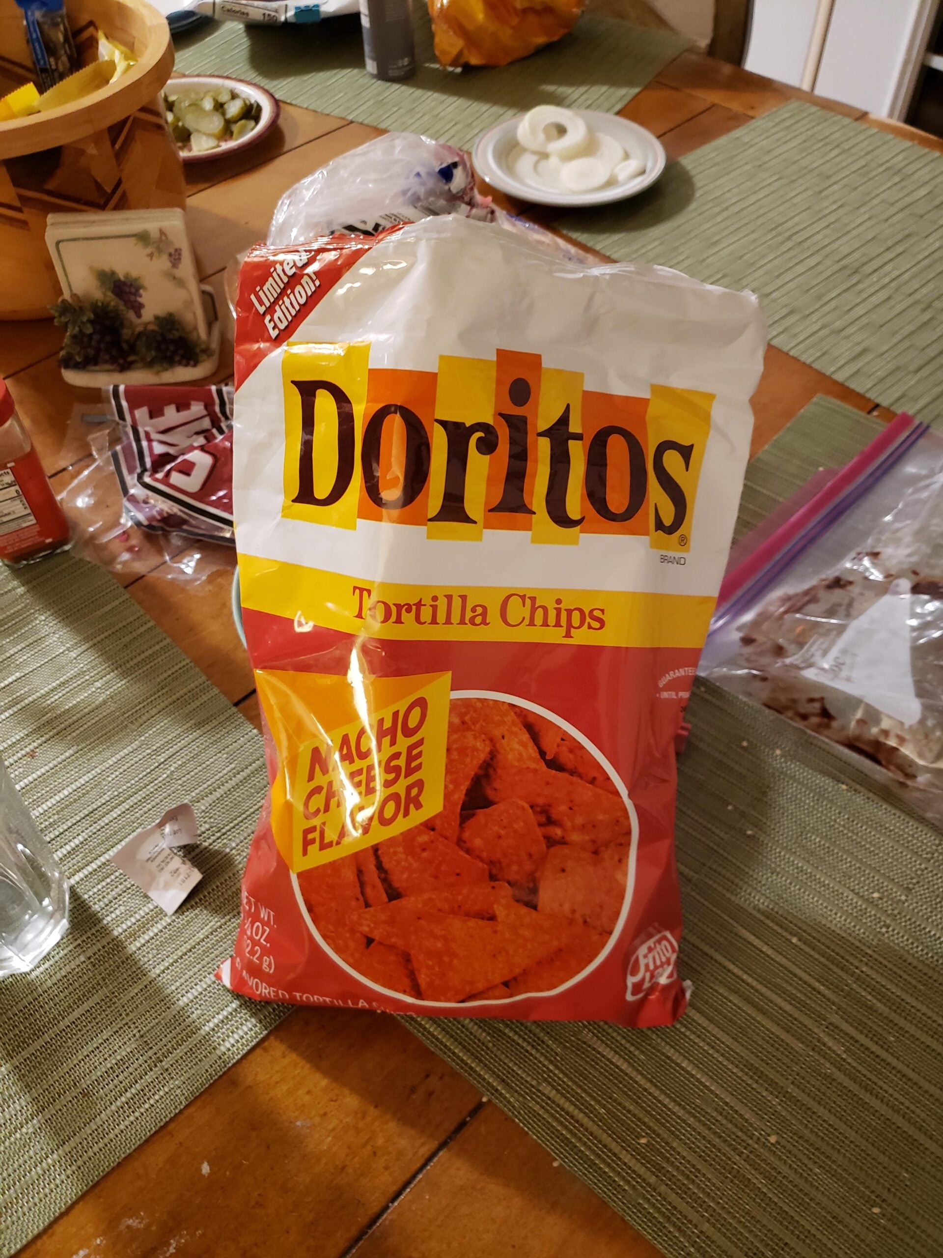 A large, retro-style bag of Doritos Tortilla Chips with Nacho Cheese flavor sits on a wooden table with a green placemat, surrounded by various food items and kitchen clutter.