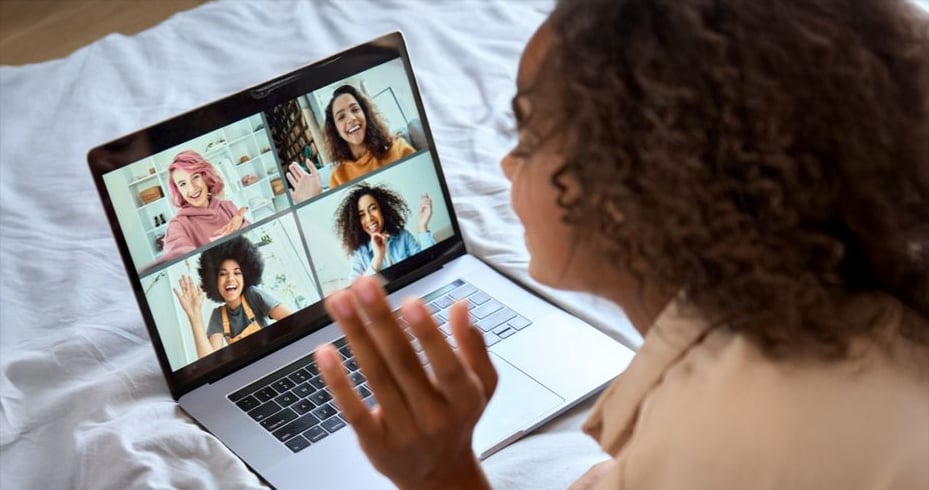 A person with curly hair sits on a bed, waving at a laptop screen showing a video call with four smiling friends, each in their own video window, greeting and chatting virtually.