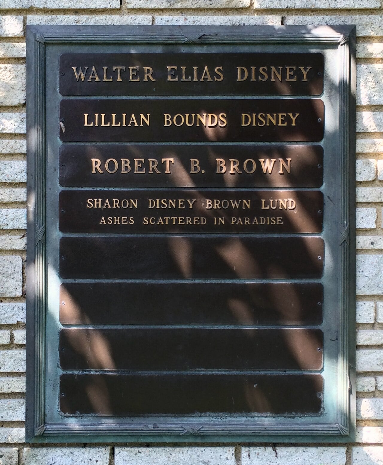 A bronze memorial plaque on a brick wall lists the names Walter Elias Disney, Lillian Bounds Disney, Robert B. Brown, and Sharon Disney Brown Lund, with a note that Sharon's ashes were scattered in paradise.