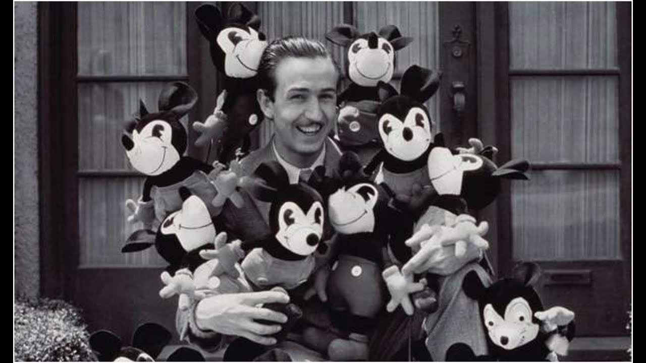 A smiling man in a suit holds a large bundle of vintage Mickey Mouse plush toys while sitting outside in front of a building with glass-paneled doors. The image is black and white.