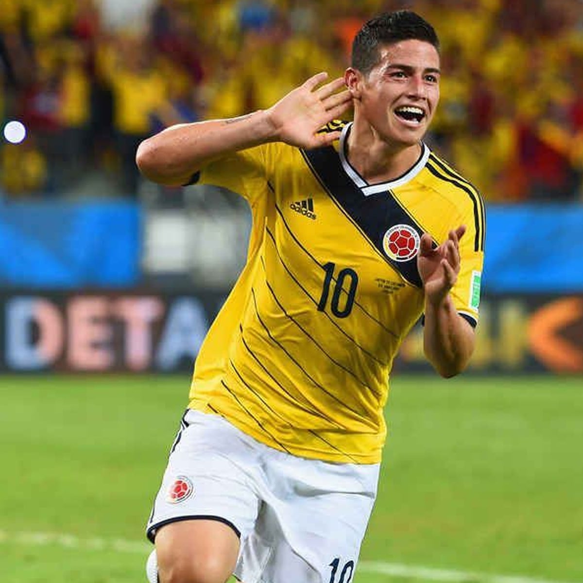 A soccer player in a yellow Colombia jersey with number 10 celebrates on the field, smiling and holding a hand to his ear, with a crowd of fans in the background.