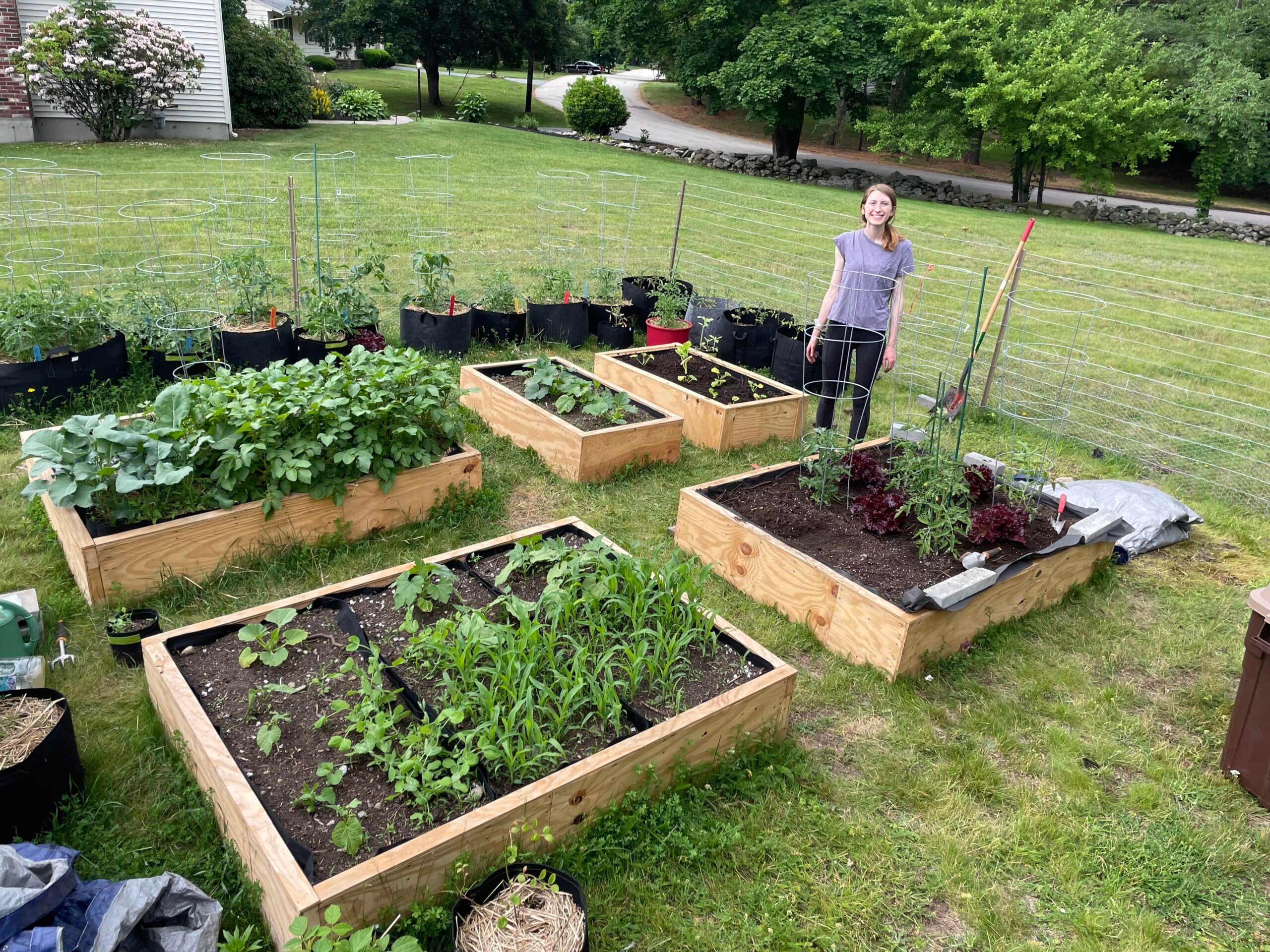 A person stands smiling in a yard next to several raised garden beds filled with various green plants and vegetables, surrounded by more planters and greenery on a cloudy day.