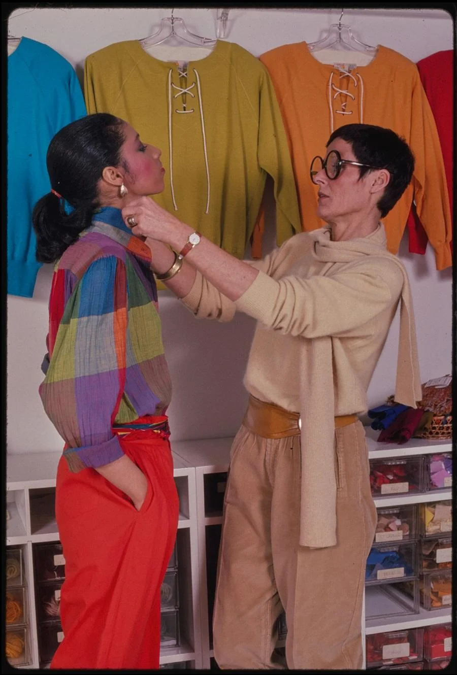 A fashion designer adjusts a model’s colorful outfit in a clothing store, with bright sweaters hanging on the wall behind them. The designer wears large glasses and a beige outfit.
