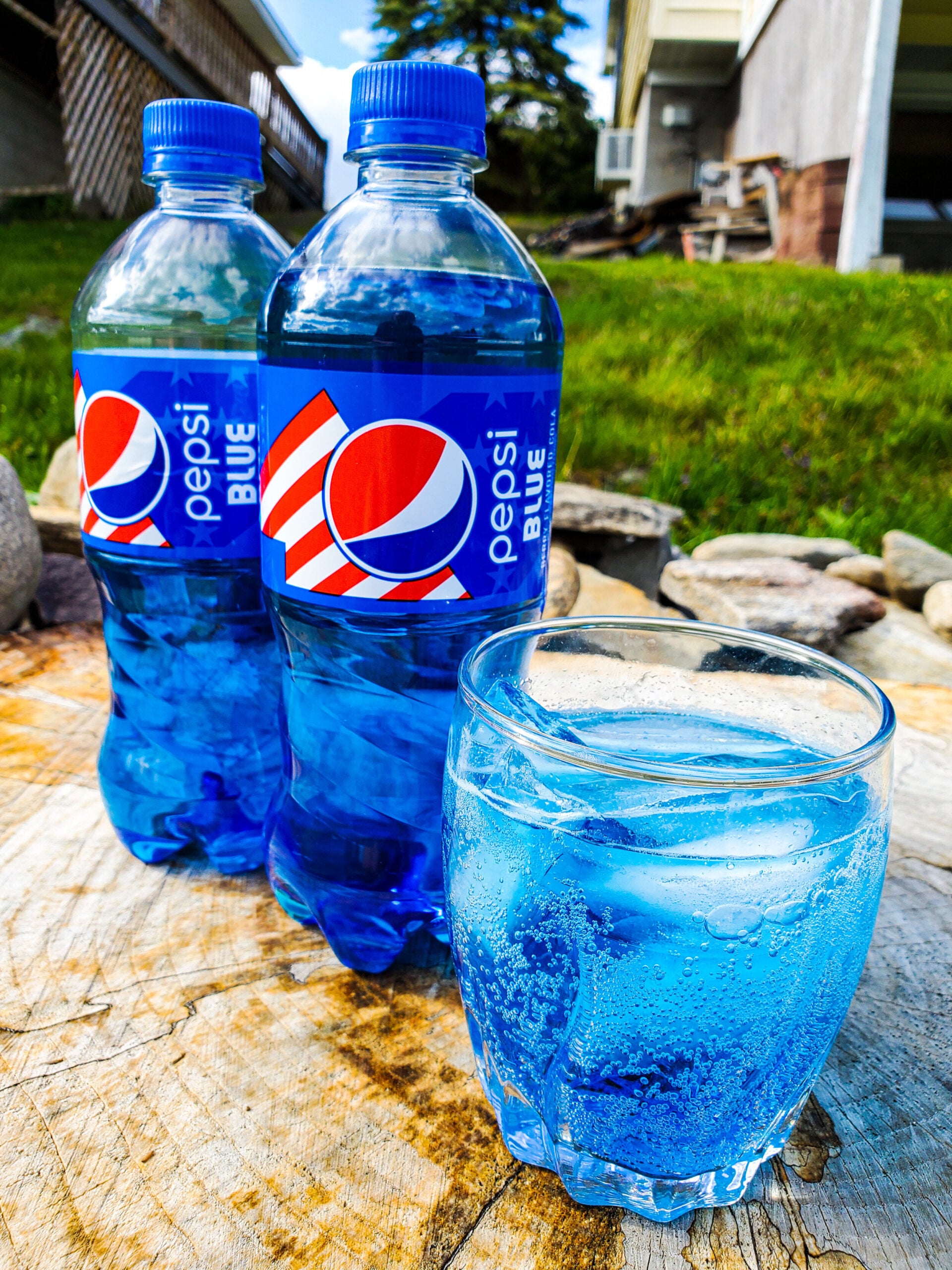 Two bottles of Pepsi Blue and a glass filled with blue soda and ice are placed on a wooden surface outdoors, with grass and a building in the background.