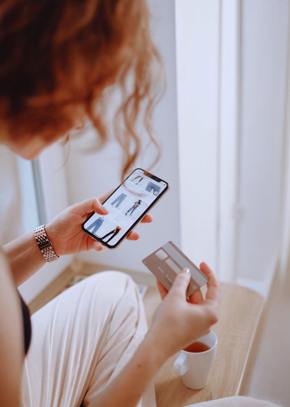 A person with curly hair browses clothing on a smartphone while holding a credit card and a white mug, sitting by a window in natural light.