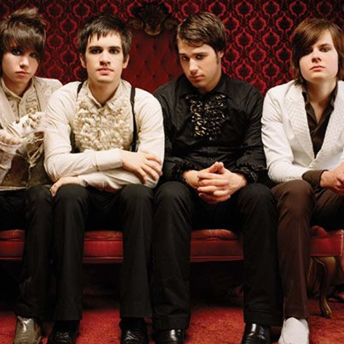 Four young men in vintage-style formal outfits sit on a red couch against a red patterned wallpaper. They have serious expressions and varying hairstyles, looking directly at the camera.