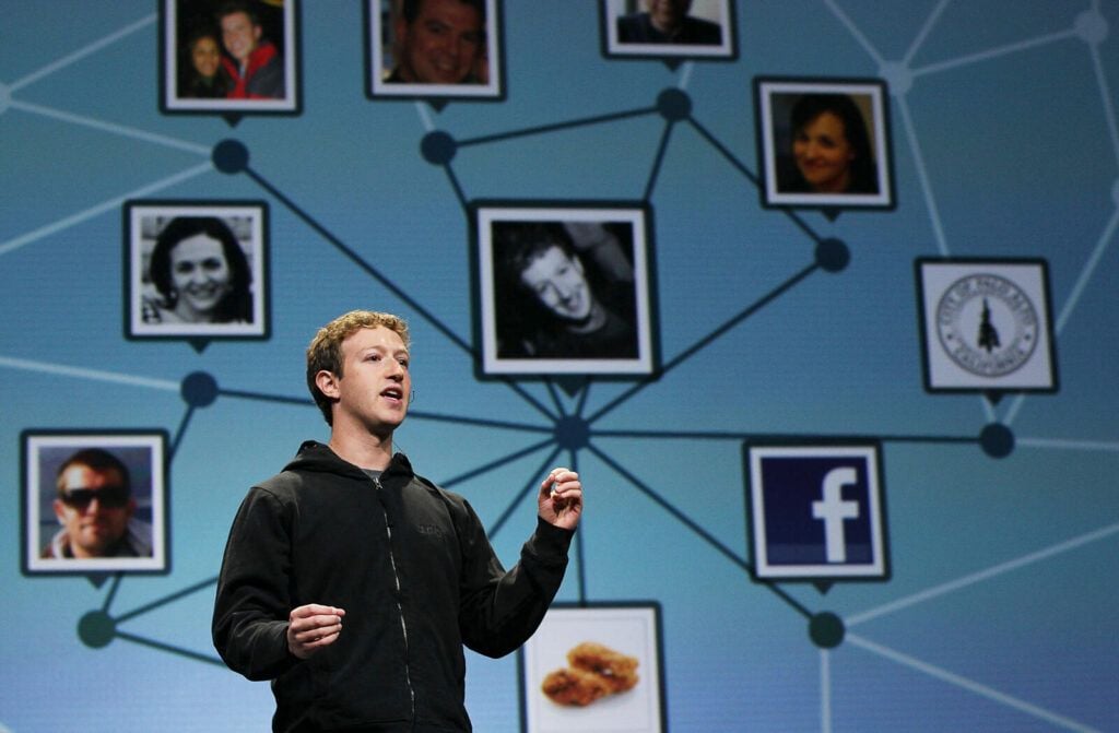 A man in a black hoodie speaks on stage in front of a screen displaying a network of profile photos, the Facebook logo, and connected nodes on a blue background.