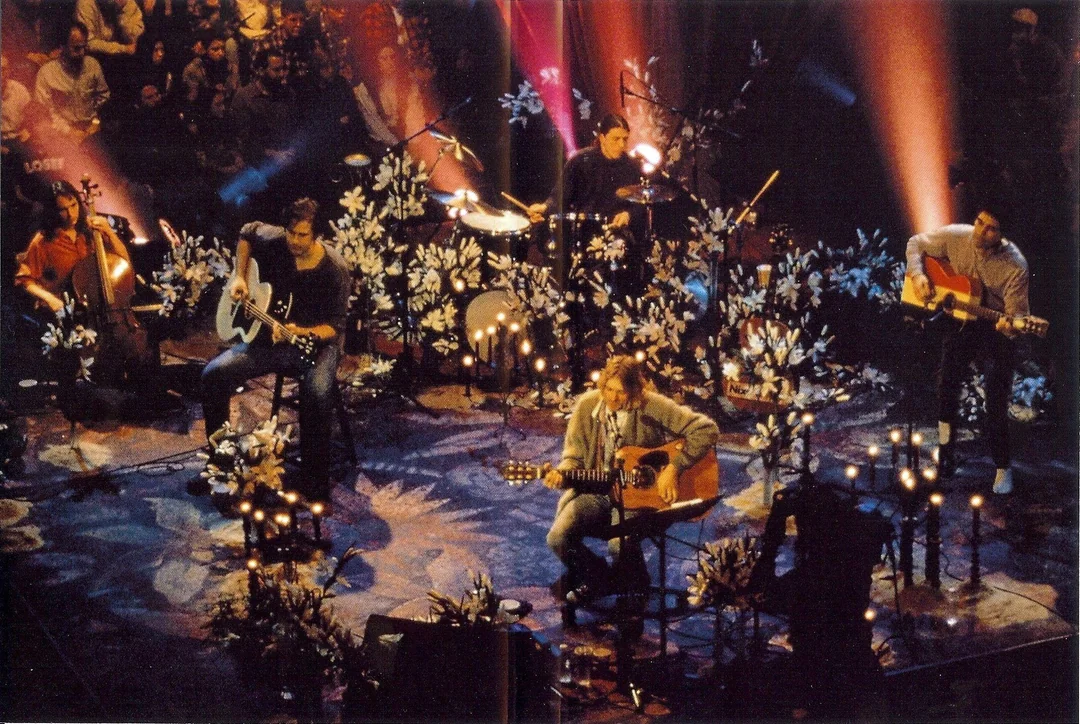 A band performs an acoustic set on a dimly lit stage decorated with white flowers and candles, surrounded by an audience. Musicians sit on stools playing guitars and drums, with warm spotlights illuminating the scene.