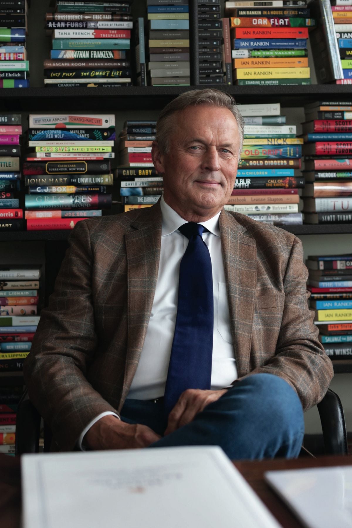 A man in a plaid blazer, white shirt, and blue tie sits in front of bookshelves filled with colorful books, looking at the camera with a slight smile. A table with documents is visible in the foreground.