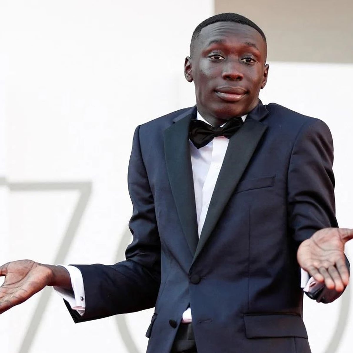 A man in a dark tuxedo with a bow tie stands with both hands raised and palms up, making a shrugging gesture and a neutral expression, as if to say, "I don't know.