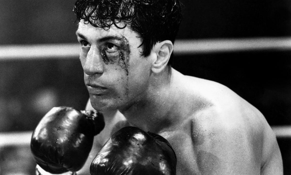 A black-and-white image of a male boxer in a ring, wearing gloves and looking upward with determination. He has a visible cut above his left eye, with blood running down his face.