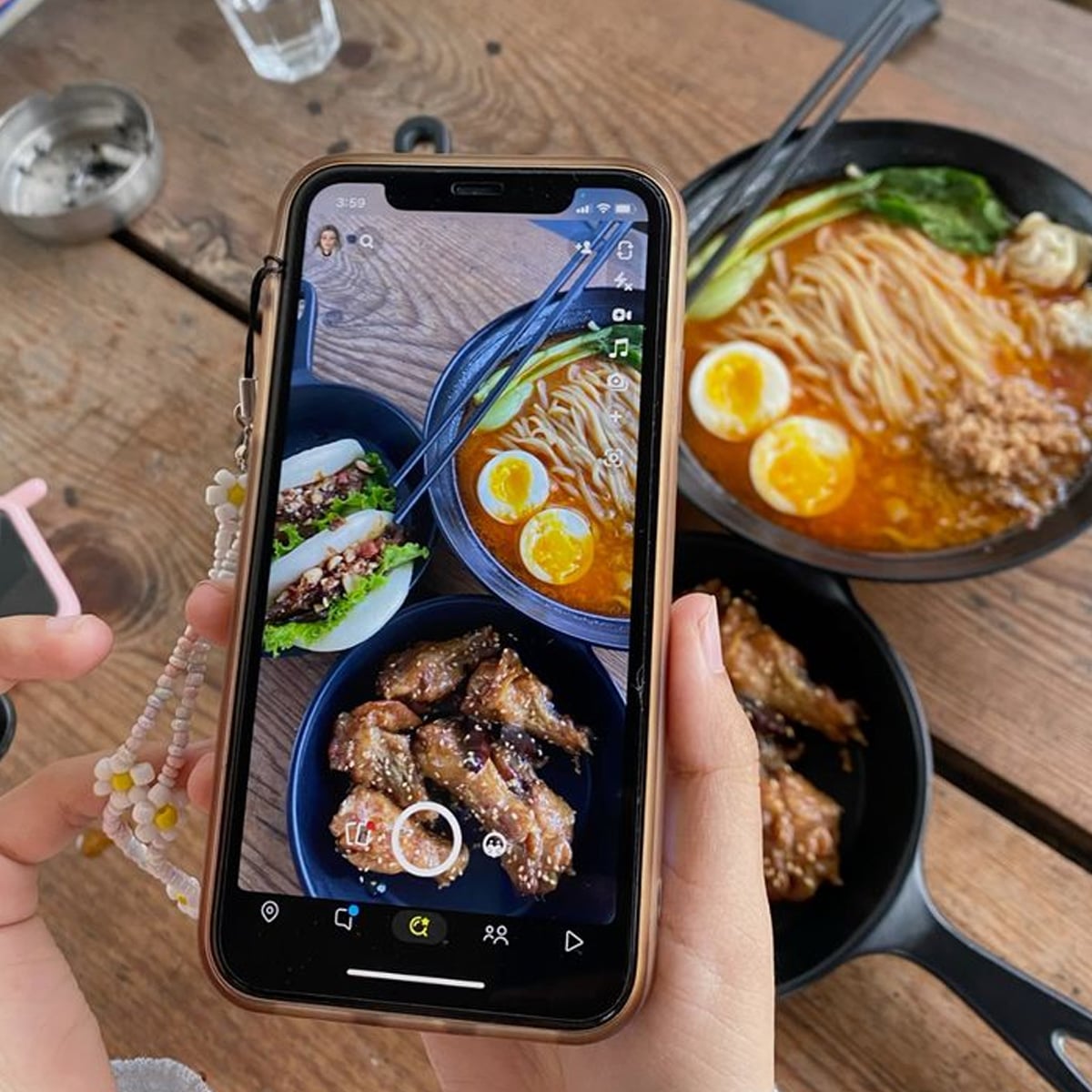 A person holds a phone taking a photo of a table with bowls of ramen, steamed buns, glazed chicken wings, and chopsticks on a wooden surface. The phone screen shows the same food from above.