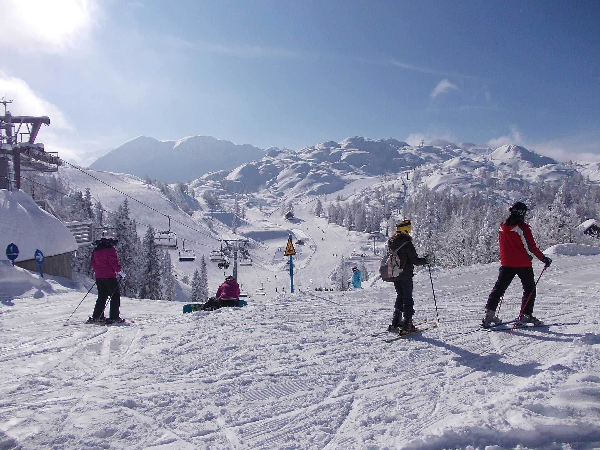 Four skiers stand and sit on a snowy slope, with ski lifts, snow-covered trees, and mountains in the background under a bright blue sky.