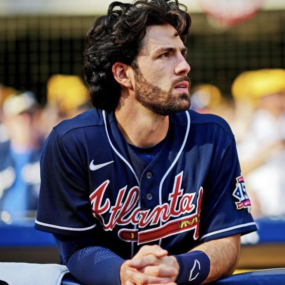 A baseball player with wavy dark hair and a beard, wearing an Atlanta Braves jersey, sits at the dugout rail and looks to the side. The stadium background is blurred with fans in the stands.