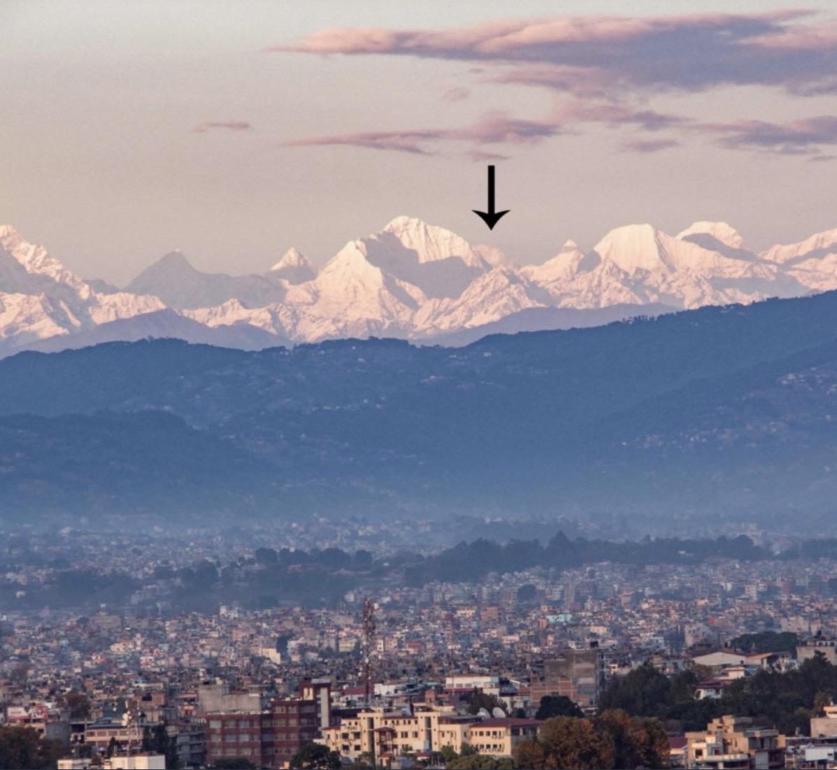 Snow-covered Himalayan mountains rise above a cityscape at dawn, with an arrow pointing to a prominent peak in the center; soft pink and purple hues fill the sky.