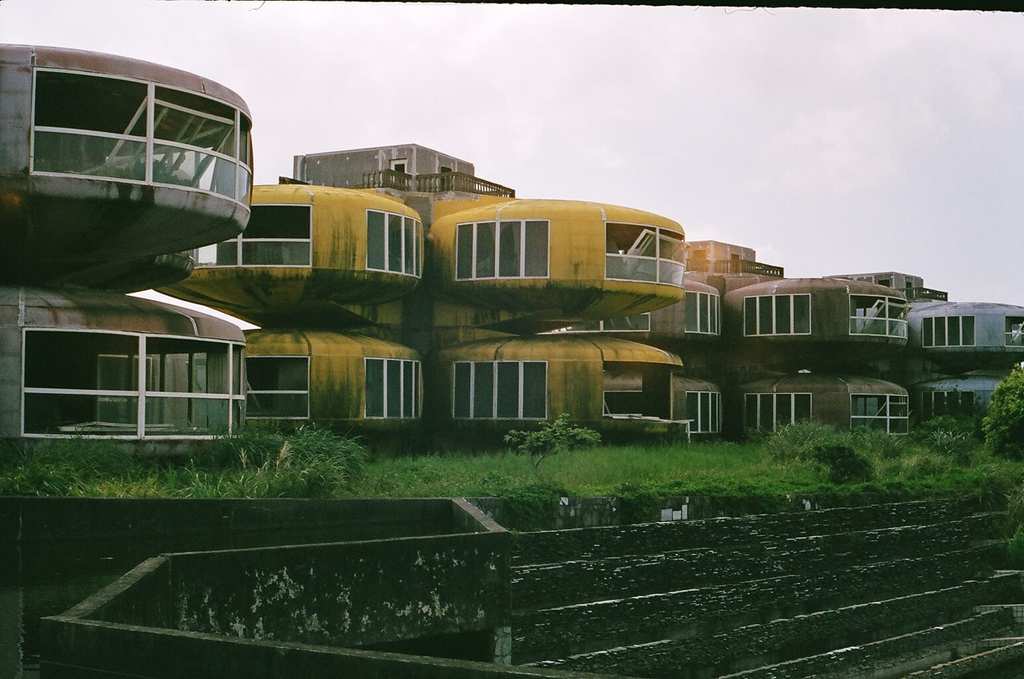 Two-story, pod-shaped buildings with large windows, some in yellow and others metallic, stand abandoned and weathered, surrounded by overgrown grass and concrete steps under a cloudy sky.