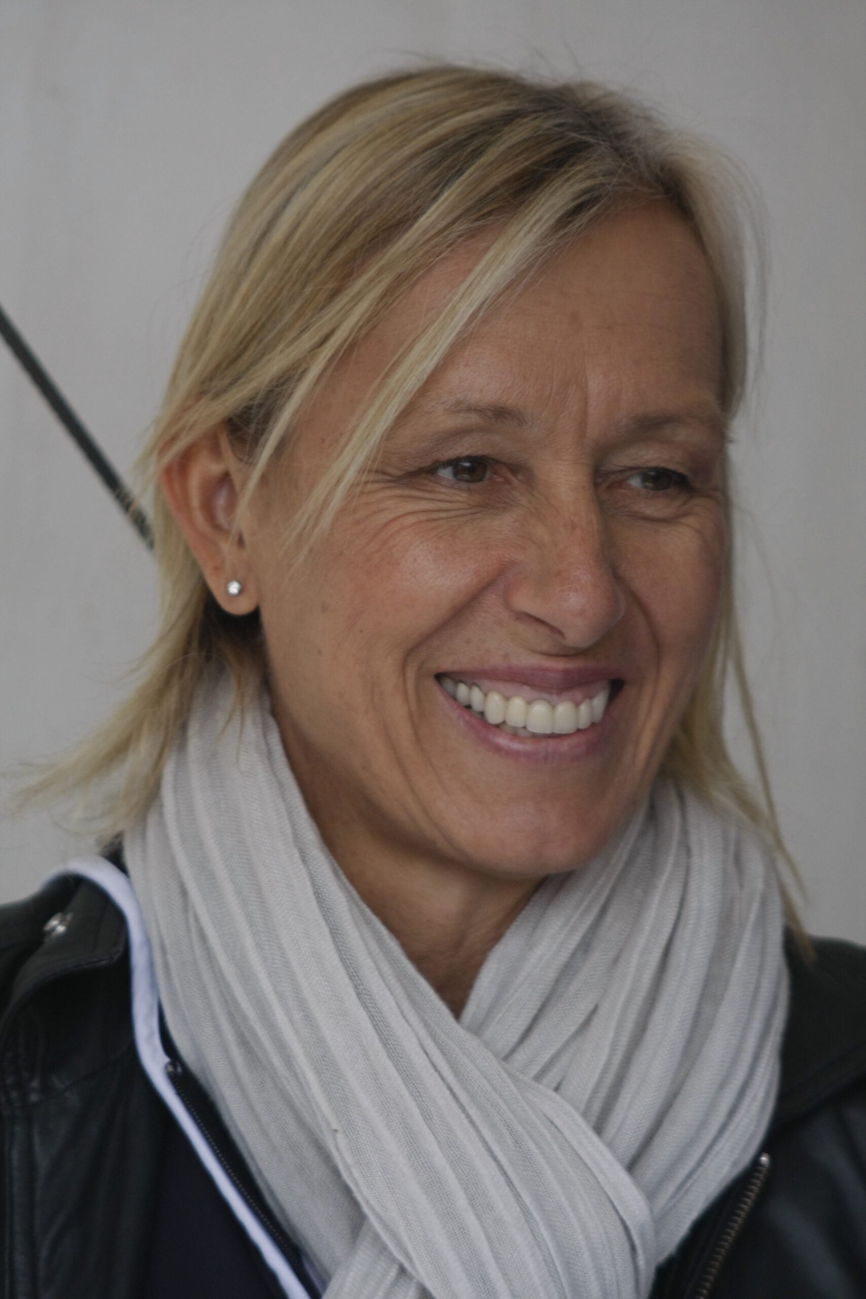 A woman with blonde hair smiles while wearing a light gray scarf, black jacket, and small earrings. The background is plain and out of focus.