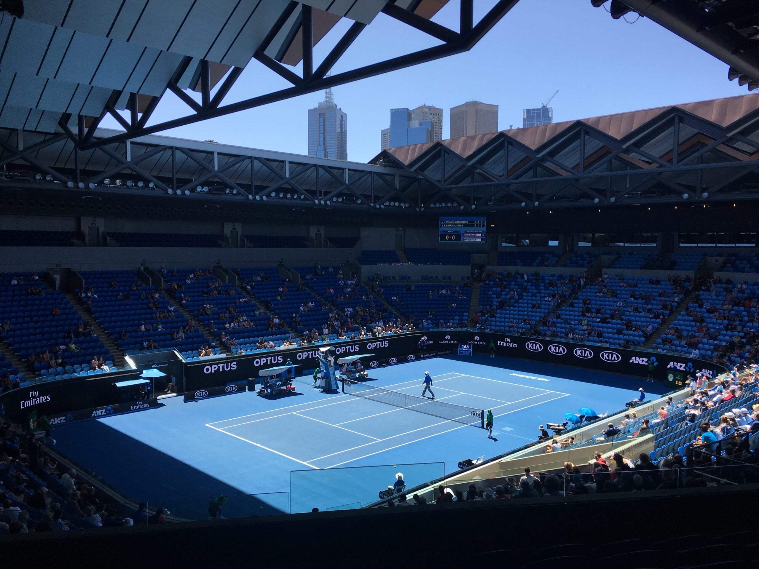A professional tennis match is underway in a large stadium with a blue court, surrounded by mostly empty blue seats. Tall city buildings are visible outside the open roof under a clear sky.