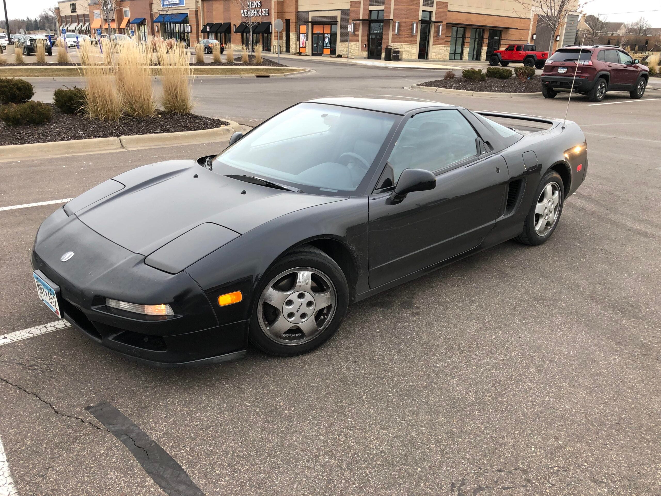 A black Acura NSX sports car is parked in a nearly empty parking lot near storefronts and landscaping with shrubs and tall grasses. The car's sleek, low design and two-door coupe style are visible.