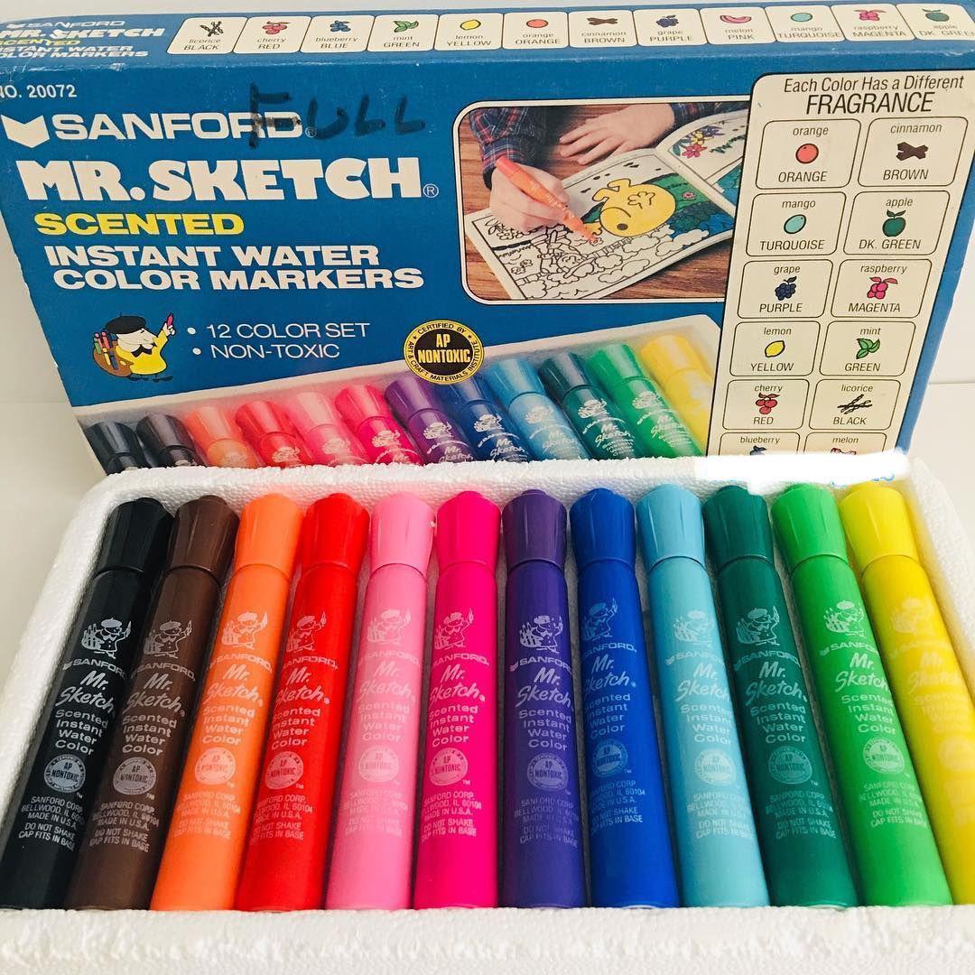 A box and tray of Mr. Sketch scented watercolor markers, showing twelve colorful markers arranged in a row below the packaging, which displays a child coloring with the markers. Each marker has a different scent.